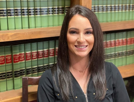 Smiling woman in a black blouse sits in a law library with shelves of green books in the background.